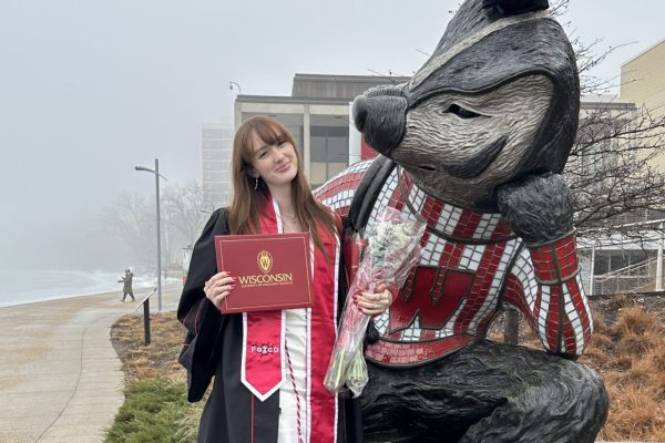 A young woman is sitting on a Bucky Badger statue while holding her diploma and several bouquets of roses