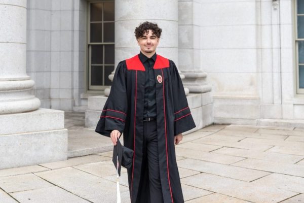 A young man walks while wearing a graduation gown and holding a graduation cap in his right hand.