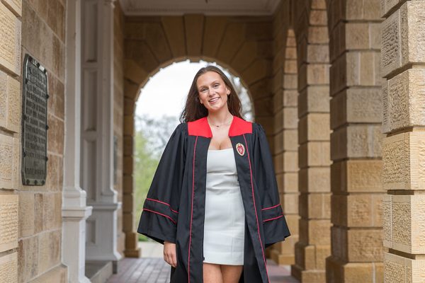 A young woman wearing a graduation gown and a white dress is walking beneath brick arches